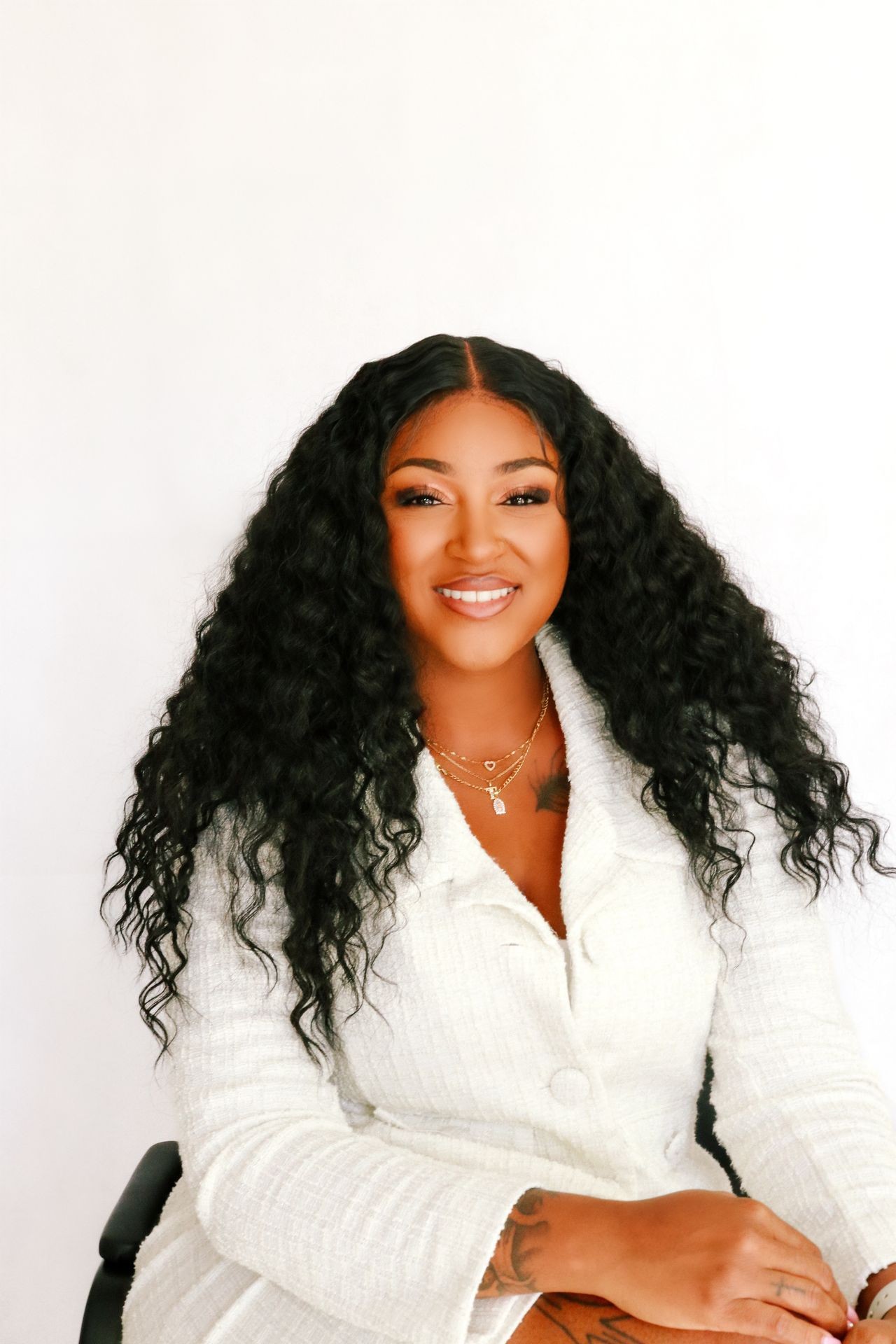 Smiling woman with curly black hair wearing a white jacket and gold necklaces against a plain background.
