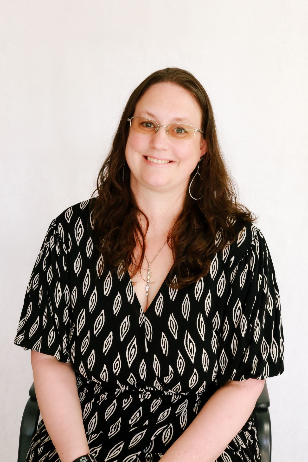 Smiling person with glasses and dark patterned dress sitting against a light background.