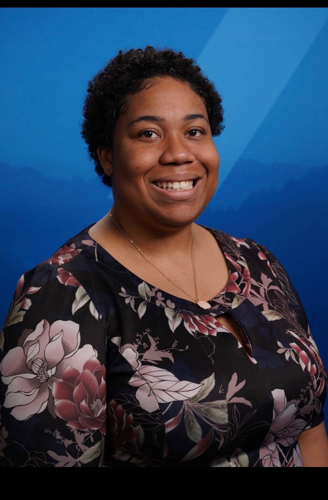 Person smiling, wearing a floral-patterned dress against a blue background.